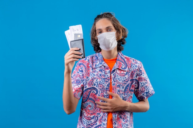 Free photo young handsome guy wearing facial protective mask holding air tickets looking with confident expression standing over blue background