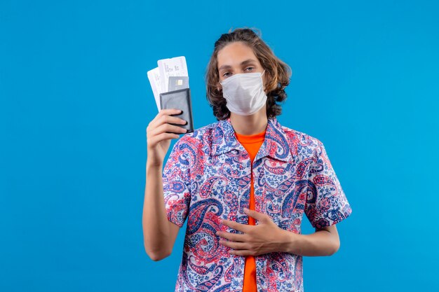 Young handsome guy wearing facial protective mask holding air tickets looking with confident expression standing over blue background