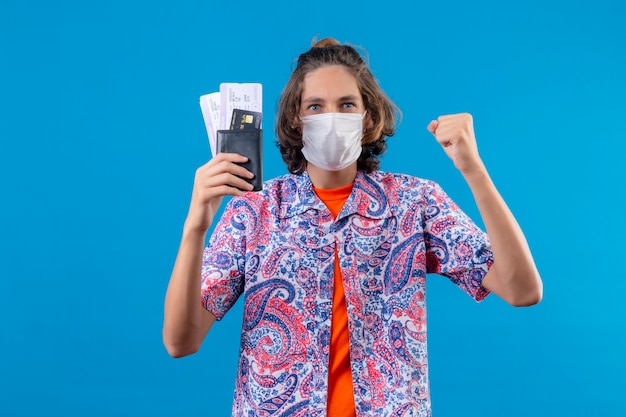 Free photo young handsome guy wearing facial protective mask holding air tickets raising fist after a victory with happy faceexited and smiling winner concept  standing over blue background