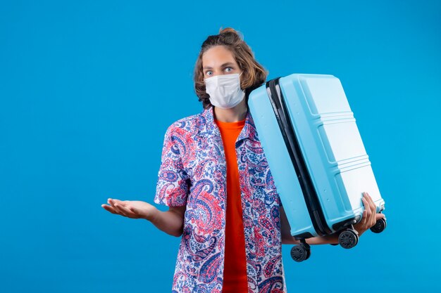 Young handsome guy wearing facial protective mask holding travel suitcase clueless and confused standing with arms raised over blue background