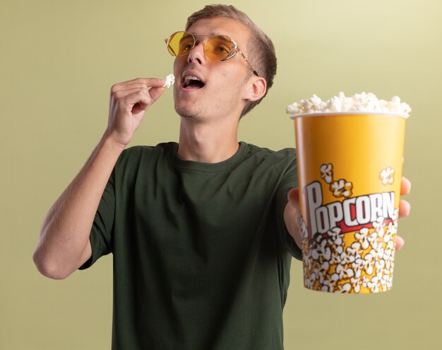 Young handsome guy wearing green shirt with glasses holding out bucket of popcorn at camera isolated on olive green wall