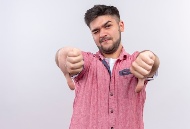 Free photo young handsome guy wearing pink polo shirt blinking en eye doing unhappy thumbs down standing over white wall