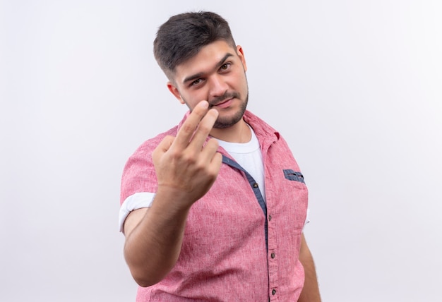 Foto gratuita giovane ragazzo bello che indossa la maglietta polo rosa facendo ti sto guardando firmare con le dita in piedi sopra il muro bianco