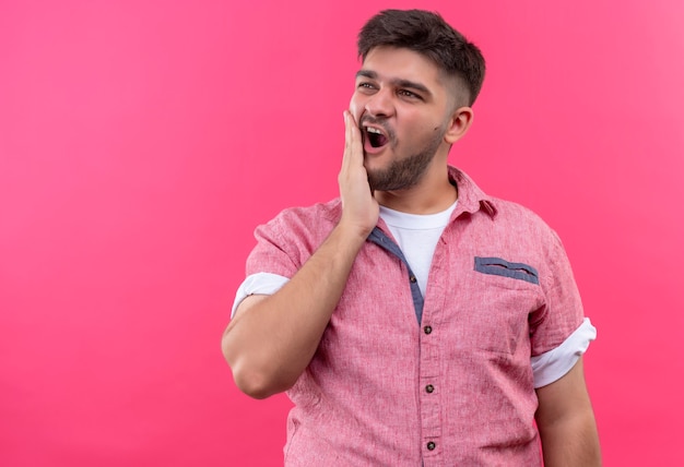 Young handsome guy wearing pink polo shirt feeling pain in teeth holding cheek with hand lokking besides standing over pink wall