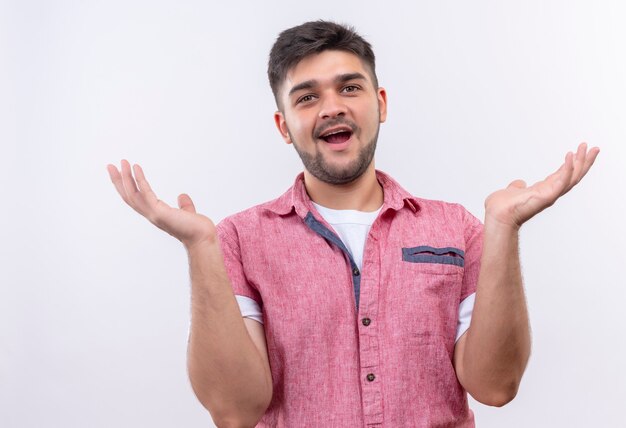 Young handsome guy wearing pink polo shirt happily looking undecided standing over white wall