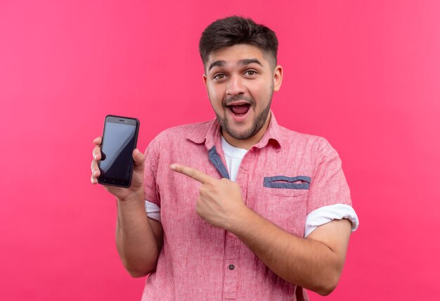 Молодой красивый парень в розовой рубашке поло, счастливо указывая на телефон с указательным пальцем, стоящим над розовой стеной