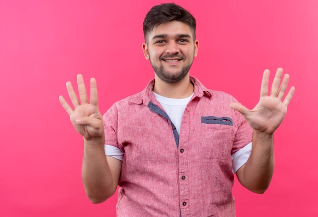 Young handsome guy wearing pink polo shirt happily showing nine sign with fingers standing over pink wall