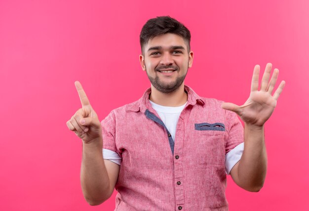 Young handsome guy wearing pink polo shirt happily showing six with fingers standing over pink wall