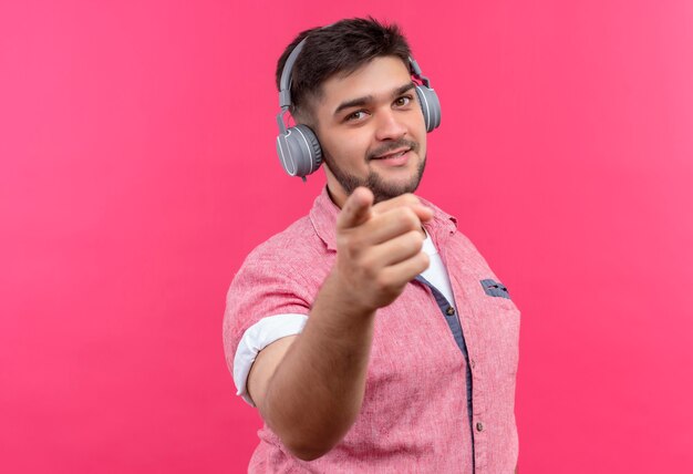 Молодой красивый парень в розовой рубашке поло и наушниках, счастливо указывая указательным пальцем на розовую стену