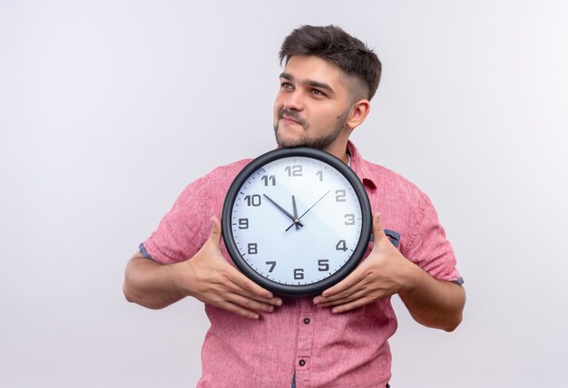 Young handsome guy wearing pink polo shirt holding clock standing over white wall