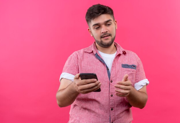 Young handsome guy wearing pink polo shirt holding phone looking down undecided standing over pink wall