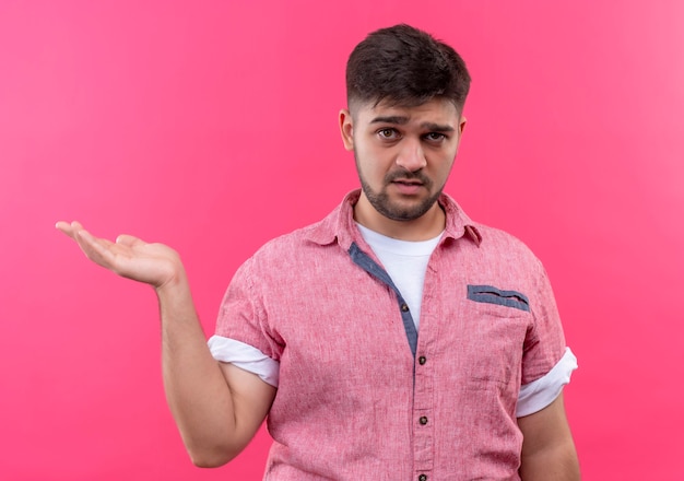 Young handsome guy wearing pink polo shirt holding right hand up confused standing over pink wall