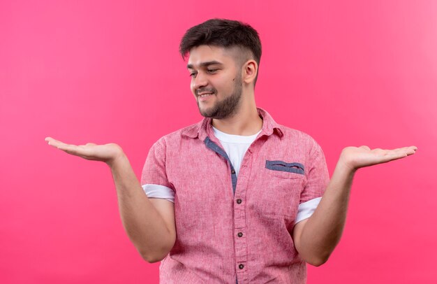 Young handsome guy wearing pink polo shirt holding something in his left and right hands looking at the right hand standing over pink wall