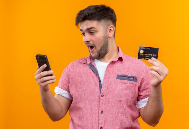 오렌지 벽 위에 서있는 신용 카드를 들고 전화를보고 분홍색 폴로 셔츠를 입고 젊은 잘 생긴 남자