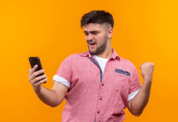 Young handsome guy wearing pink polo shirt looking at phone, standing over orange wall