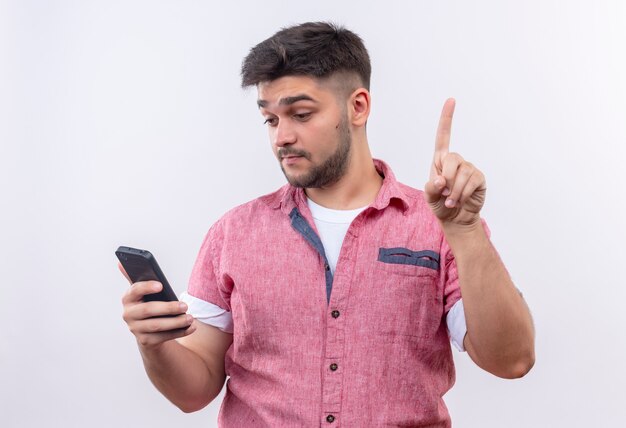 Young handsome guy wearing pink polo shirt looking at phone thoughtfully doing up his forefinger standing over white wall