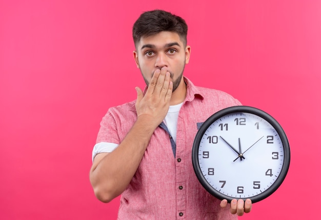 Young handsome guy wearing pink polo shirt looking shocked holding clock afraid of being late standing over pink wall