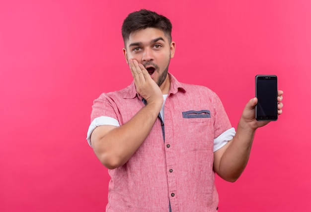 Young handsome guy wearing pink polo shirt looking shocked holding phone standing over pink wall