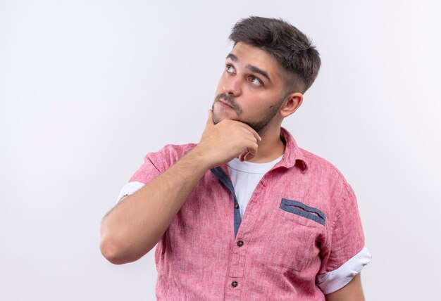 Young handsome guy wearing pink polo shirt looking thoughtfully up standing over white wall