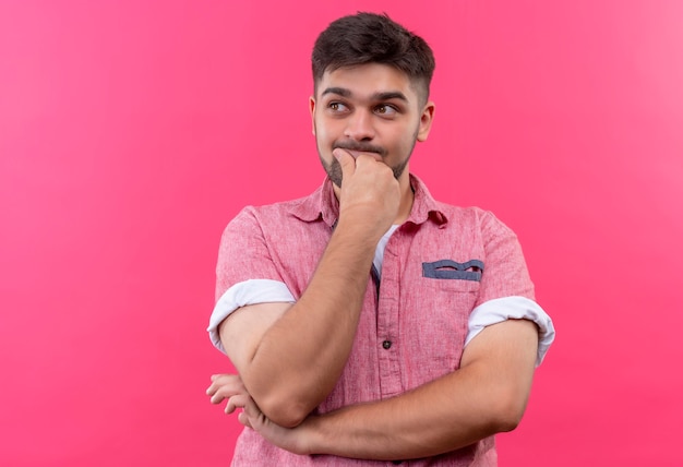 Giovane ragazzo bello che indossa la maglietta polo rosa che sembra indeciso oltre a stare sopra il muro rosa