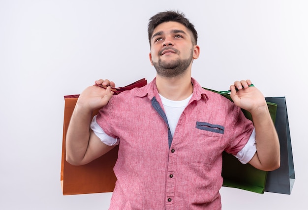 Young handsome guy wearing pink polo shirt looking up pleased holding shopping bags in his back standing over white wall