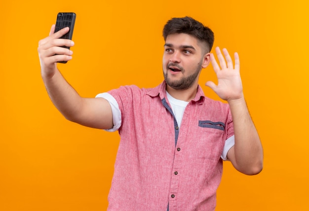 Young handsome guy wearing pink polo shirt making selfie saying hi standing over orange wall