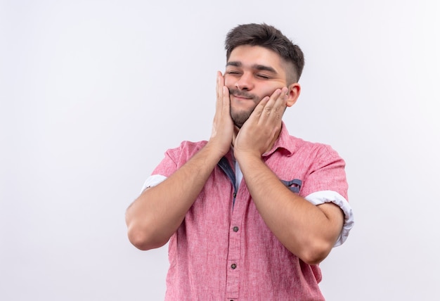 Young handsome guy wearing pink polo shirt massaging his face standing over white wall
