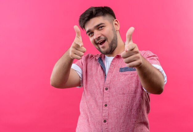 Молодой красивый парень в розовой рубашке поло, игриво указывая указательными пальцами на розовую стену
