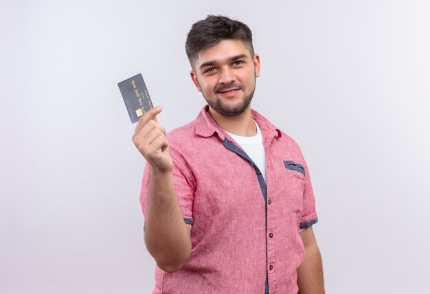 ピンクのポロシャツを着た若いハンサムな男は、白い壁の上に立っているクレジットカードを持って見て喜んでいます