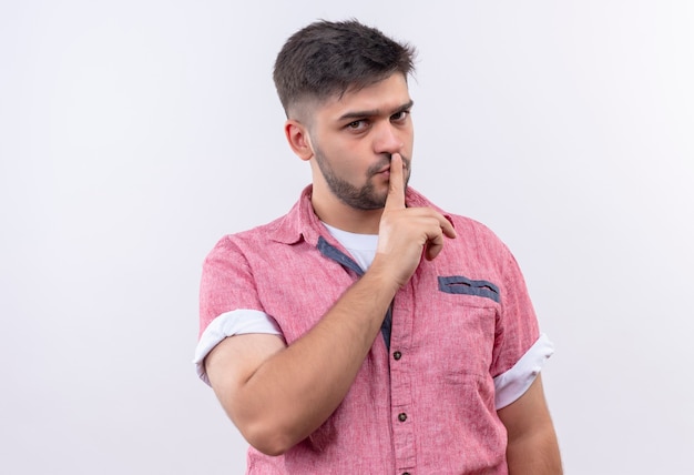 Giovane ragazzo bello che indossa la maglietta polo rosa con rimprovero facendo segno di silenzio alla telecamera con l'indice in piedi sopra il muro bianco