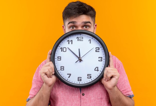 Young handsome guy wearing pink polo shirt scared hiding behind clock for being late standing over orange wall