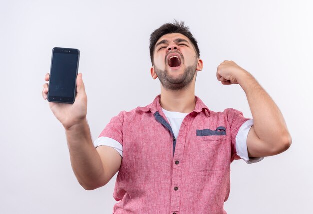 Young handsome guy wearing pink polo shirt shaking his fist holding phone closing eyes standing over white wall