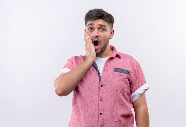 Young handsome guy wearing pink polo shirt shocked looking holding his cheek with the left hand standing over white wall