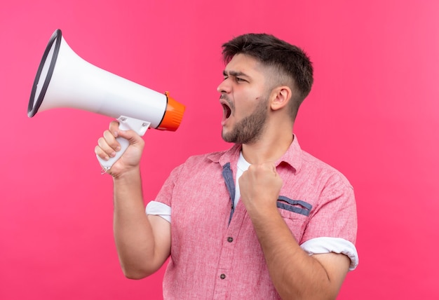 Free photo young handsome guy wearing pink polo shirt shouting on megaphone standing over pink wall