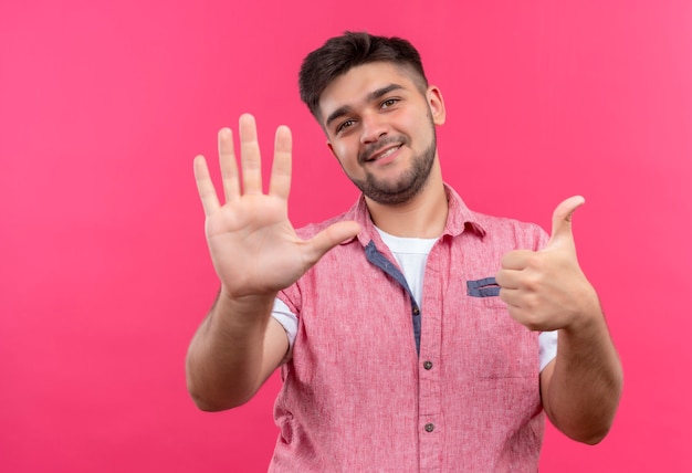 Foto gratuita giovane ragazzo bello che indossa la maglietta polo rosa che mostra cinque e uno in piedi sul muro rosa