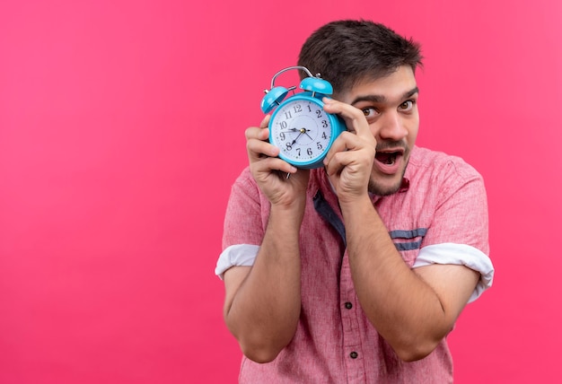 Young handsome guy wearing pink polo shirt showing that it's time holding blue alarm clock standing over pink wall