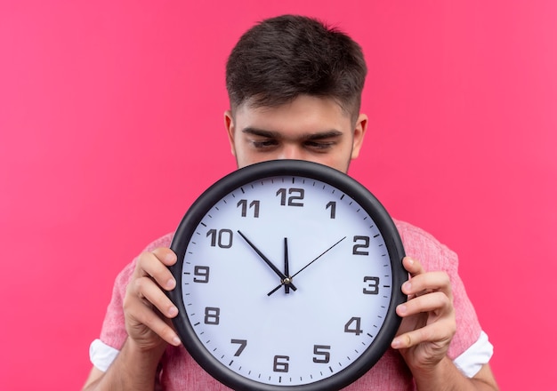Young handsome guy wearing pink polo shirt sleepy holding clock tanding over pink wall