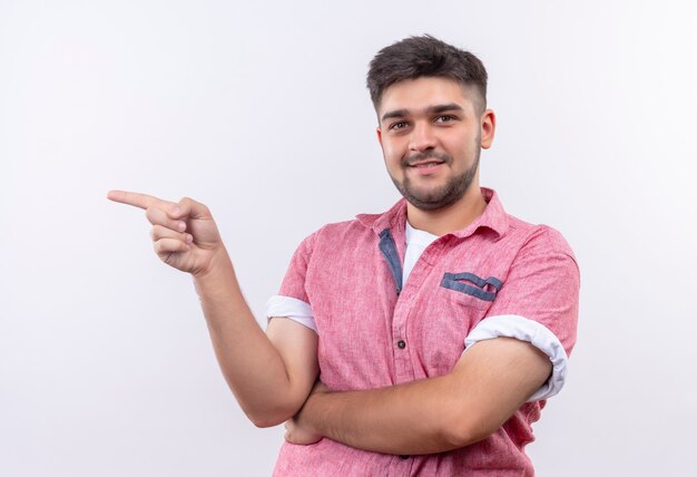 Young handsome guy wearing pink polo shirt smiling coridally pointing to the right with forefinger standing over white wall
