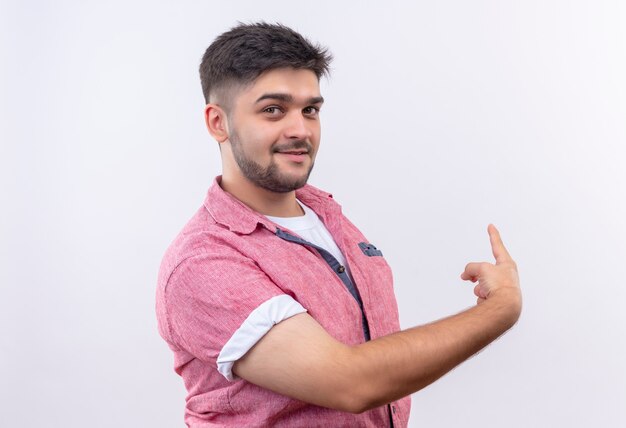 Young handsome guy wearing pink polo shirt smiling showing back with forefinger standing over white wall