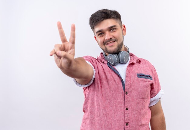 Young handsome guy wearing pink polo shirt smiling showing peace sign with fingers standing over white wall