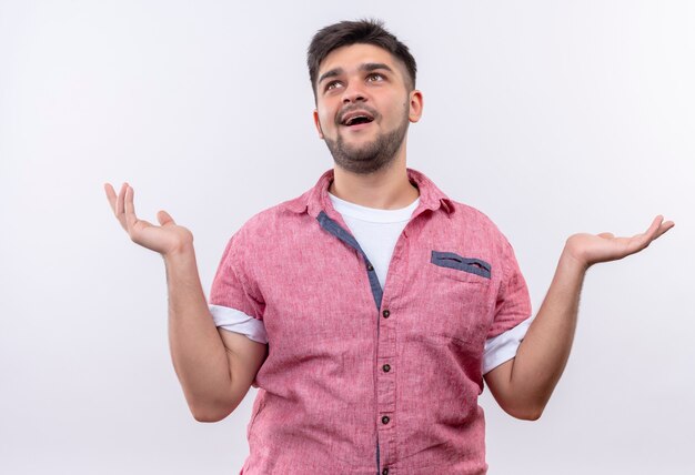 Young handsome guy wearing pink polo shirt smiling up undecided standing over white wall