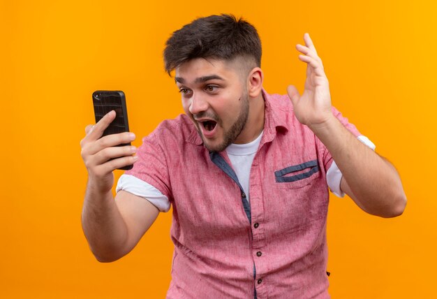 Young handsome guy wearing pink polo shirt surpised looking at phone standing over orange wall