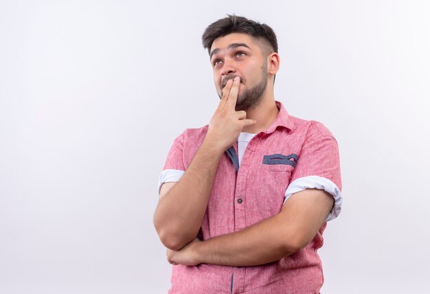 Young handsome guy wearing pink polo shirt thoughtfully looking up touching mouth with fingers standing over white wall