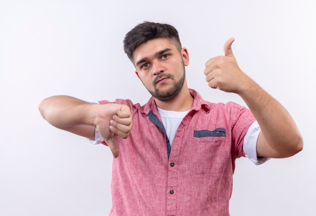 Young handsome guy wearing pink polo shirt undecided looking shwoing happy and unhappy thumbs standing over white wall