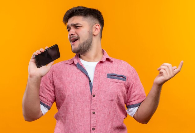 Young handsome guy wearing pink polo singing playfully on phone shirt standing over orange wall