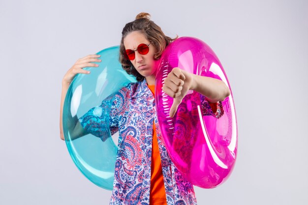 Young handsome guy wearing red sunglasses holding colorful inflatable rings displeased showing thumbs down standing over white background