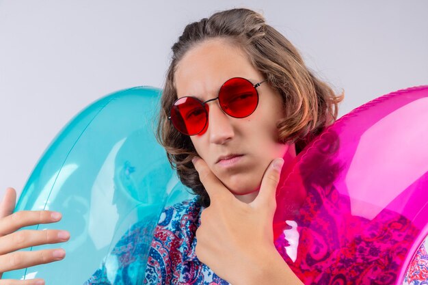 Young handsome guy wearing red sunglasses holding colorful inflatable rings looking at camera with pensive suspicious expression with hand on chin thinking standing over white background