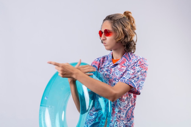 Young handsome guy wearing red sunglasses holding inflatable ring looking aside with confused expression pointing to something standing 
