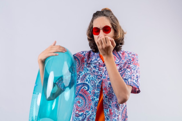 Young handsome guy wearing red sunglasses holding  inflatable ring looking stressed and nervous biting nails standing over white background