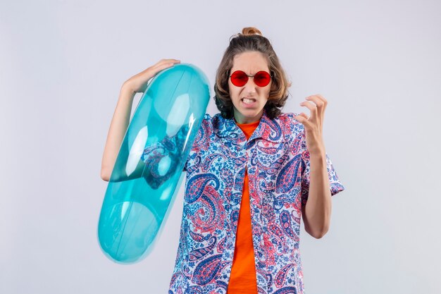 Young handsome guy wearing red sunglasses holding  inflatable ring mad and crazy standing with arms raised over white background